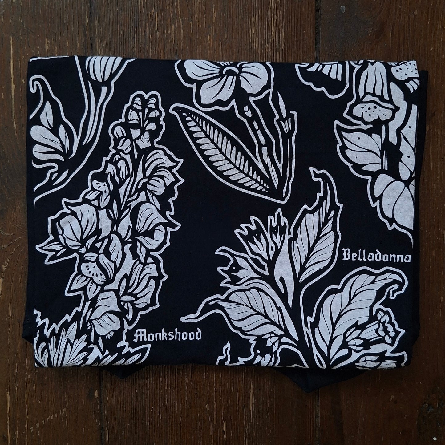 A folded black tehirt featuring a white botanical block print with the words 'Belladonna' and 'Monkshood' printed on it.