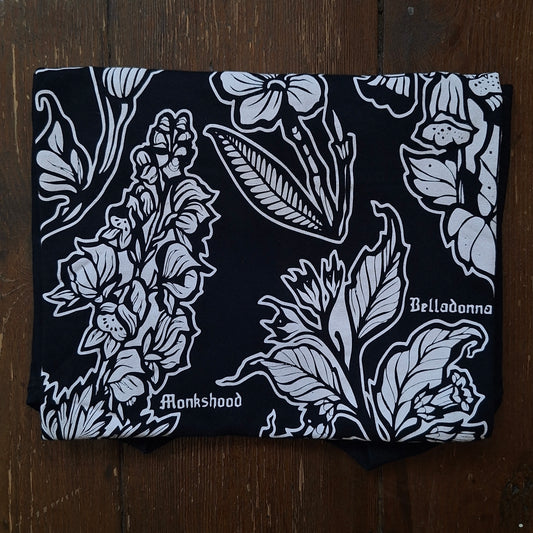 A folded black tehirt featuring a white botanical block print with the words 'Belladonna' and 'Monkshood' printed on it.